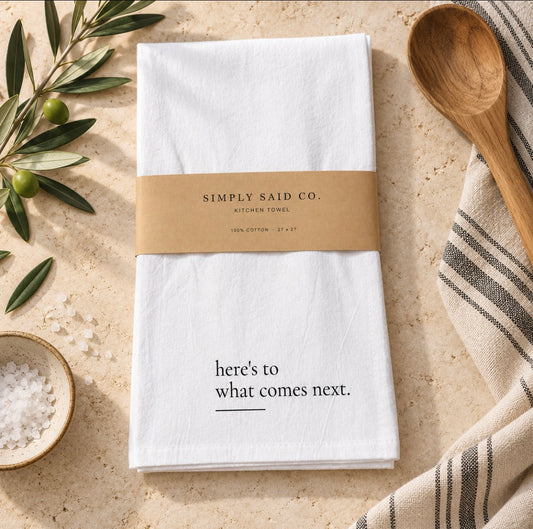 A white cotton kitchen towel with black text reading 'here's to what comes next.' is wrapped in a kraft paper band labeled 'SIMPLY SAID CO.
