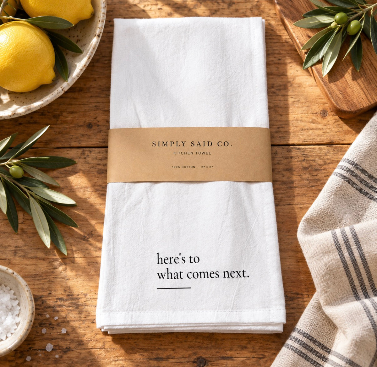Here’s to What Comes Next quote kitchen towel styled on rustic wood with lemons