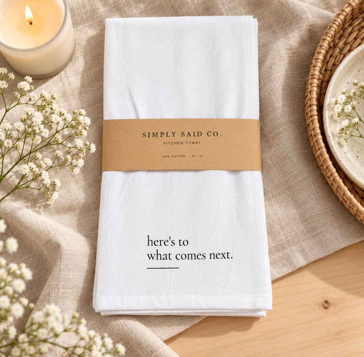 A folded white kitchen towel with black printed text reading 'here's to what comes next.' sits on a beige linen cloth, wrapped with a brown paper band that says 'SIMPLY SAID CO.