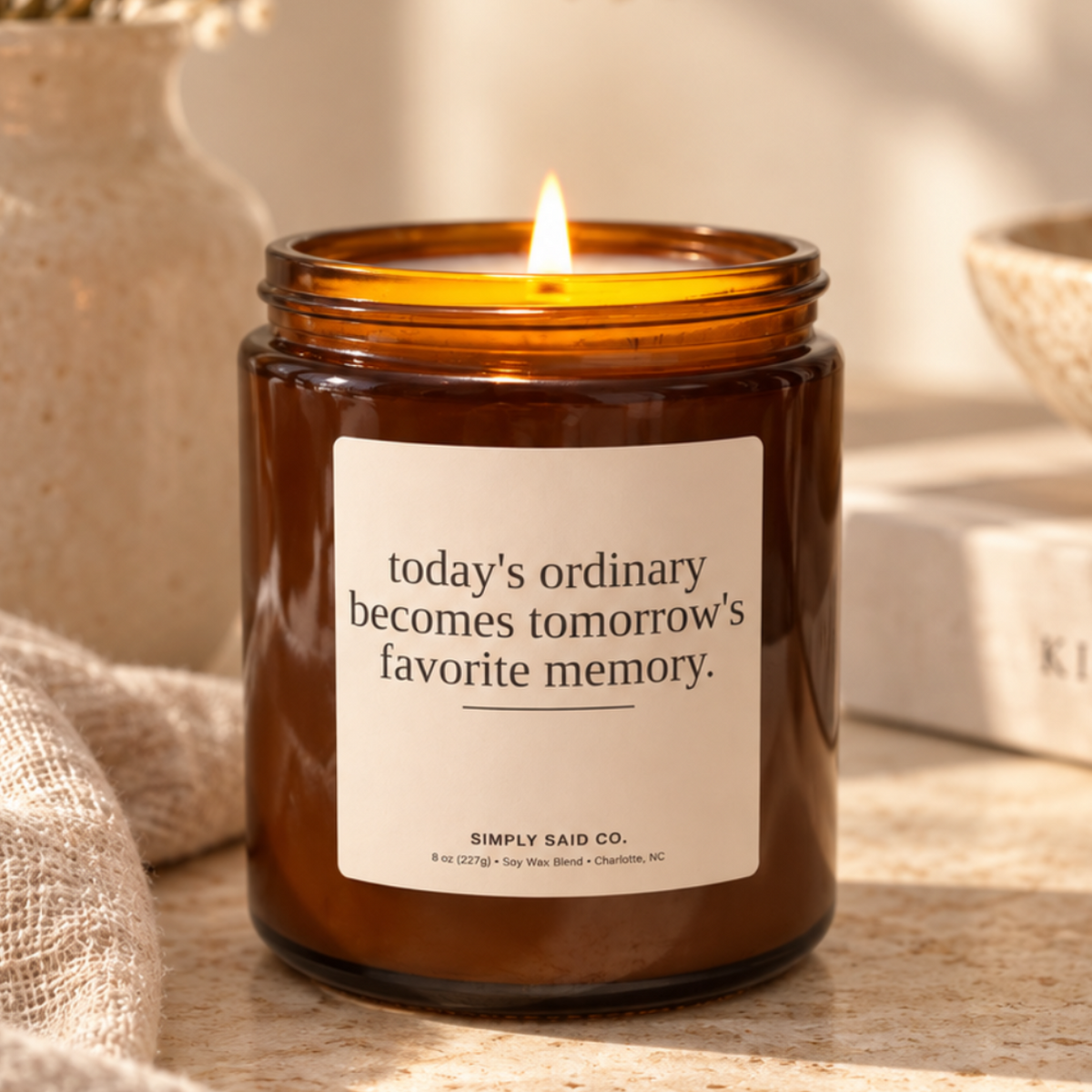 A lit amber glass jar candle sits on a stone surface with a linen cloth, dried flowers in a vase, and a book in a warm styled setting. The label reads, "today's ordinary becomes tomorrow's favorite memory.
